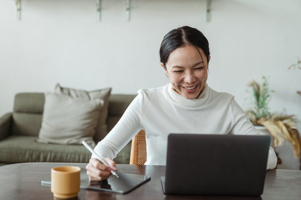 A cheerful woman uses a laptop and tablet for a video call, working remotely in a cozy living room.